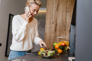 Mutfakta sağlıklı salata yaparken cep telefonu kullanan güzel, gülümseyen genç bir kadın.