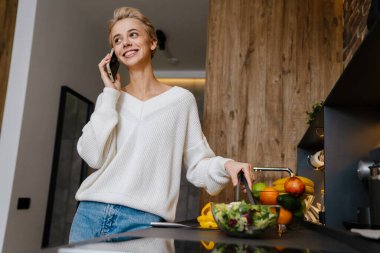 Mutfakta sağlıklı salata yaparken cep telefonu kullanan güzel, gülümseyen genç bir kadın.