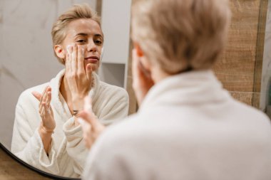 Çekici genç bir kadın banyoda dururken yüzüne nemlendirici sürüyor.