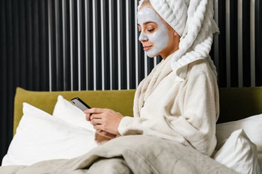 Yatakta otururken cep telefonu kullanan, bornoz giyen gülümseyen genç bir kadın.