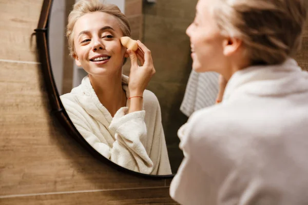 Atractiva Joven Mujer Usando Albornoz Aplicando Maquillaje Con Esponja Mientras — Foto de Stock