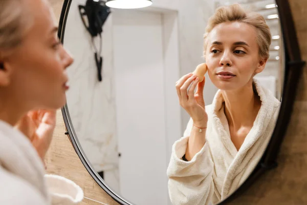 Atractiva Joven Mujer Usando Albornoz Aplicando Maquillaje Con Esponja Mientras — Foto de Stock