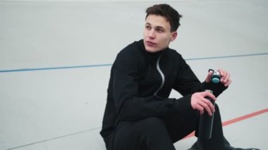 Memnun bir genç sporcu spor sahasında otururken bir spor şişesinden su içiyor.