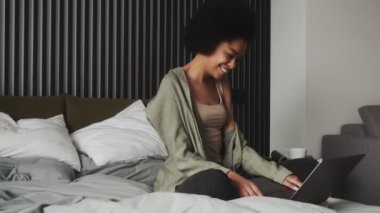 Ayaklarında dizüstü bilgisayarla odada oturmuş bir şeyler yazan siyah bir kadının yan görüntüsü.