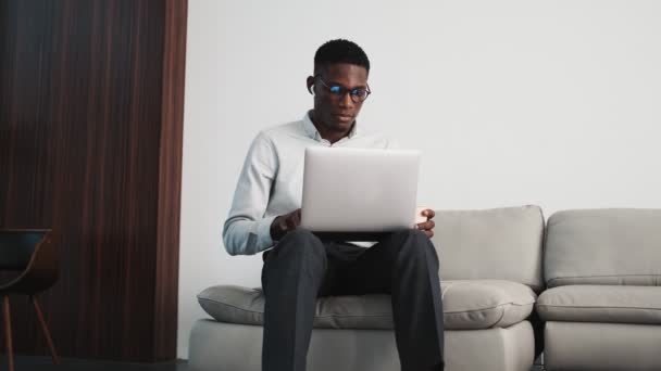 一位戴着耳塞的严肃的非洲裔美国商人正坐在公寓里的沙发上 用他的笔记本电脑把耳塞放在膝头上 — 图库视频影像