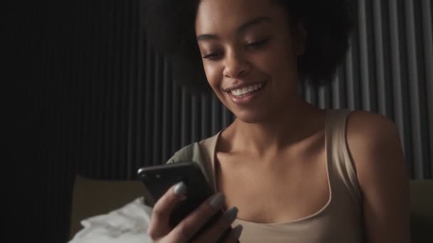 Черная Улыбающаяся Женщина Смотрит Телефон Сидя Кровати Комнате — стоковое видео