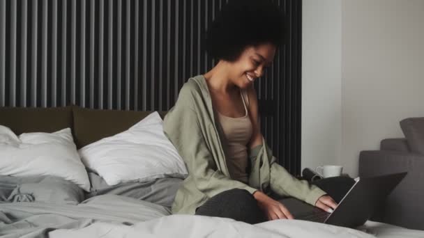 一个面带微笑的黑人女人站在手提电脑旁边 边看边在房间里的床上打字 — 图库视频影像