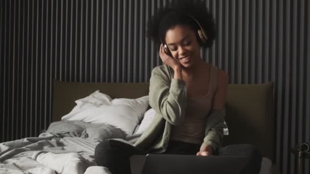 ソファの上にノートパソコンの前に座り 部屋のイヤホンの近くで手を取りながら ヘッドフォンで音楽を聴いている笑顔の女性 — ストック動画