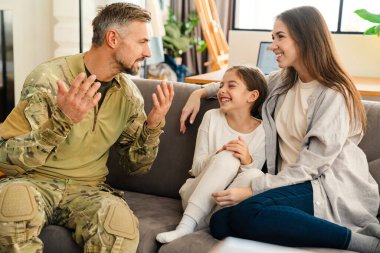 Erkeksi asker, mutlu ailesiyle evde konuşurken el kol hareketi yapıyor.