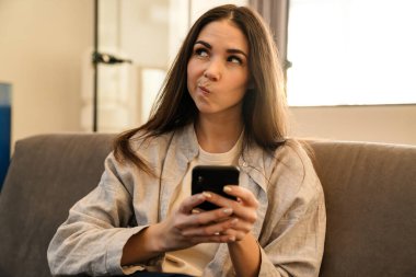 Kanepede otururken akıllı telefon kullanan kafası karışmış genç bir kadın.