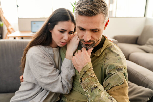 Sad young military soldier saying goodbye to sad wife at home indoors