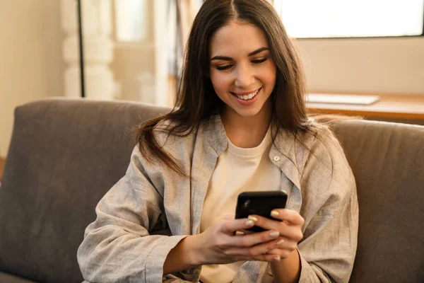 Jovem Feliz Sorrindo Usando Smartphone Enquanto Senta Sofá Dentro Casa — Fotografia de Stock