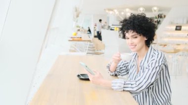 Gülümseyen bir kadın kafede oturup kahve içerken akıllı telefonundan bir şey izliyor.