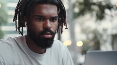 Kulaklıklı, odaklanmış bir Afrikalı Amerikalı sokakta otururken laptopunu kullanıyor.