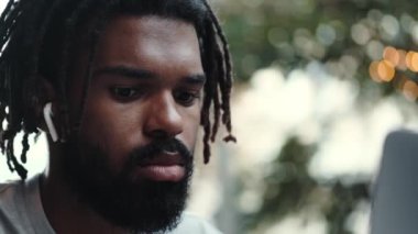 Kulaklıklı, odaklanmış bir Afrikalı Amerikalı 'nın yakından çekilmiş görüntüsü sokakta otururken dizüstü bilgisayarını kullanıyor.