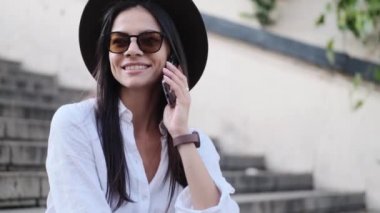 Şapka takan gülümseyen bir kadın yazın merdivenlerde oturmuş akıllı telefonuyla konuşuyor.