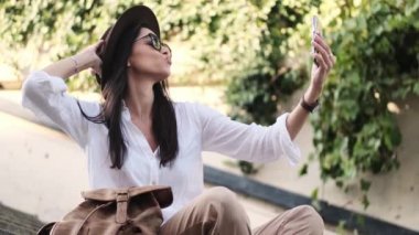 Şapka takan gülümseyen bir kadın yazın merdivenlerde oturmuş akıllı telefonunu kullanarak selfie çekiyor.