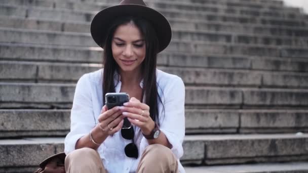 Гарна Модна Жінка Капелюсі Використовує Свій Мобільний Телефон Сидячи Сходах — стокове відео