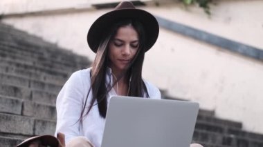 Şapka takan memnun genç bir kadın dizüstü bilgisayarını kullanarak müziği dinliyor. Yazın merdivenlerde oturuyor.