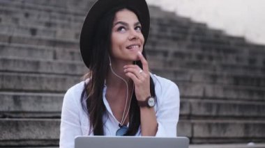 Şapka takan genç bir kadın dizüstü bilgisayarını kullanarak müziği dinliyor. Yazın merdivenlerde oturuyor.