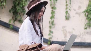 Şapka takan memnun genç bir bayanın yan görüntüsü yazın merdivenlerde oturan dizüstü bilgisayarını kullanıyor.
