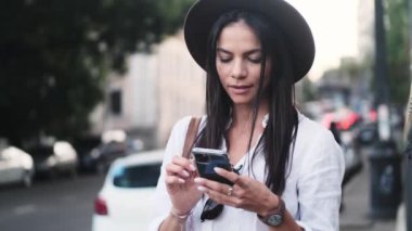 Güzel bir genç kadın yazın dışarıda caddenin kenarında dururken akıllı telefonunu kullanıyor.