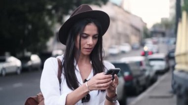 Güzel bir genç kadın yazın dışarıda caddenin kenarında dururken akıllı telefonunu kullanıyor.