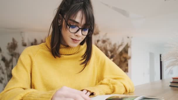 一个戴眼镜沉思的女人 穿着一件黄色的毛衣 翻阅一本杂志 坐在房间里看着窗外 — 图库视频影像