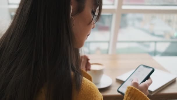 携帯電話で購入を選択し カフェのテーブルに座っている間にテープを残してヘッドフォンで女性のバックビュー — ストック動画