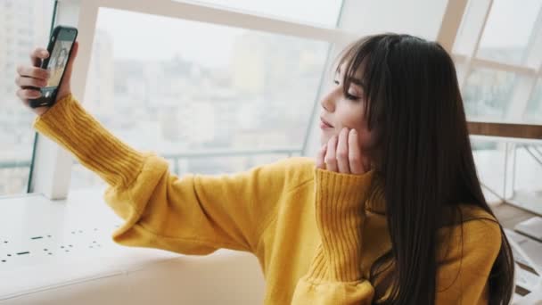 一个穿着黄色毛衣的女人坐在窗前 拿着手机自拍 摆出姿势的侧视图 — 图库视频影像