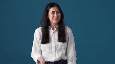 Kafası karışmış Koreli bir kadın, stüdyoda mavi duvarın üzerinde tek başına dururken, el hareketi gösteriyor.