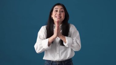Gülümseyen Koreli bir kadın, stüdyodaki mavi duvarın üzerinde el ele tutuşurken bir şey soruyor.