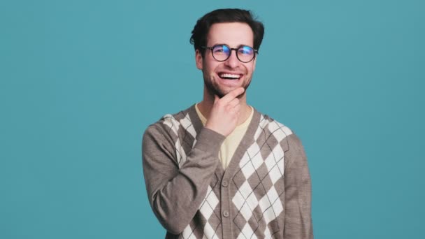 一个戴眼镜 面带微笑的男人一边想着什么 一边用手抚摩着他的胡子 同时用食指指着蓝色工作室里的相机 — 图库视频影像