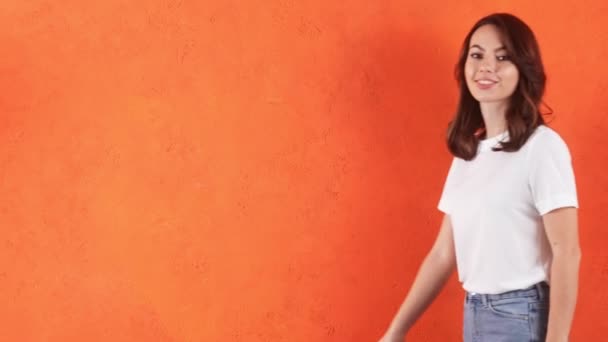 笑顔の女性が角から出てきてカメラに向かってオレンジのスタジオで手を振る — ストック動画