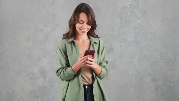 Mujer Sonriente Mirando Teléfono Mientras Sostiene Sus Manos Estudio Gris — Vídeos de Stock