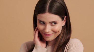 Güzel, utangaç bir genç kadının yakın plan görüntüsü stüdyodaki bej bir arka planda izole bir şekilde kameraya bakıyor.