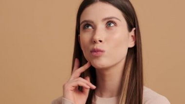 Güzel genç bir kadının yakın plan görüntüsü stüdyodaki bej bir arka planda izole edilmiş bir şeyi düşünüyor.