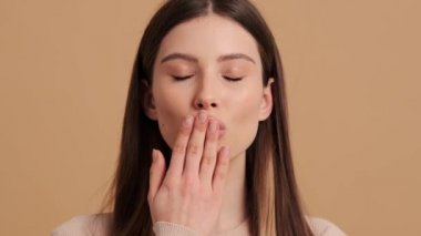 Güzel bir genç bayanın yakın çekim görüntüsü stüdyodaki bej bir arka planda izole bir şekilde kameraya hava öpücüğü gönderiyor.