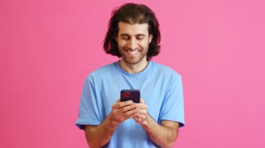 Uzun saçlı gülümseyen adam stüdyoda dururken telefonla mesaj atıyor.