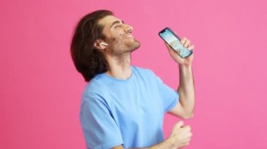 Kulaklıklı mutlu adam pembe stüdyoda elinde telefonla dans ediyor.