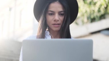 Şapka takan güzel bir genç bayanın yakın çekim görüntüsü laptopunu kullanarak yazın dışarıda merdivenlerde oturuyor.