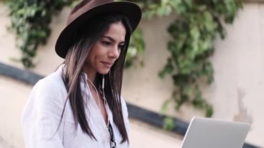 Şapka takan genç bir bayanın yan görüntüsü laptopunu kullanarak yazın dışarıdaki merdivenlerde oturuyor.