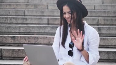 Telli kulaklık takan mutlu genç bir kadın yazın merdivenlerde oturan dizüstü bilgisayarını kullanarak video bağlantısıyla konuşurken elini sallıyor.
