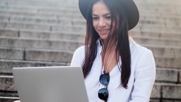 一位戴耳机的年轻貌美女子的近照是夏天坐在外面楼梯上的手提电脑 — 图库视频影像