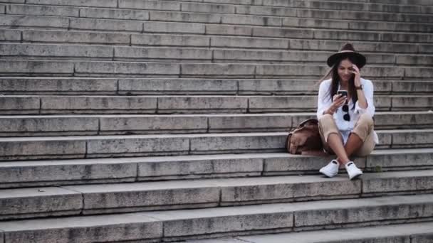 Позитивна Молода Жінка Капелюсі Використовує Свій Мобільний Телефон Сидячи Сходах — стокове відео