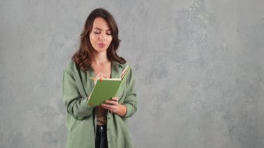 Gülümseyen bir kadın gri bir stüdyoda dururken not defterine bir şeyler yazıyor.