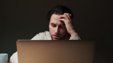 Keyifsiz yorgun bir adam dizüstü bilgisayarında çalışıyor ve gece vakti masada oturuyor.