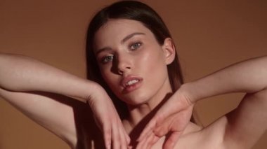 Güzel, genç, yarı çıplak bir kadın kameraya poz verirken ellerini yüzünün ve boynunun yakınına koyuyor. Stüdyoda bej bir arka planda tek başına duruyor.