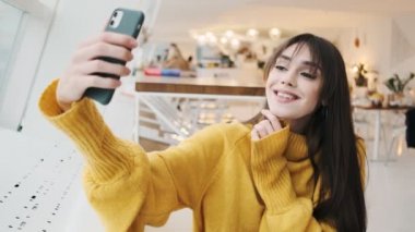 The woman in a yellow sweater taking selfie on a phone and posing with gestures while sitting in front of a window in a cafe