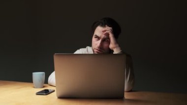 A serious focused man is working his laptop computer while sitting at the table at night time